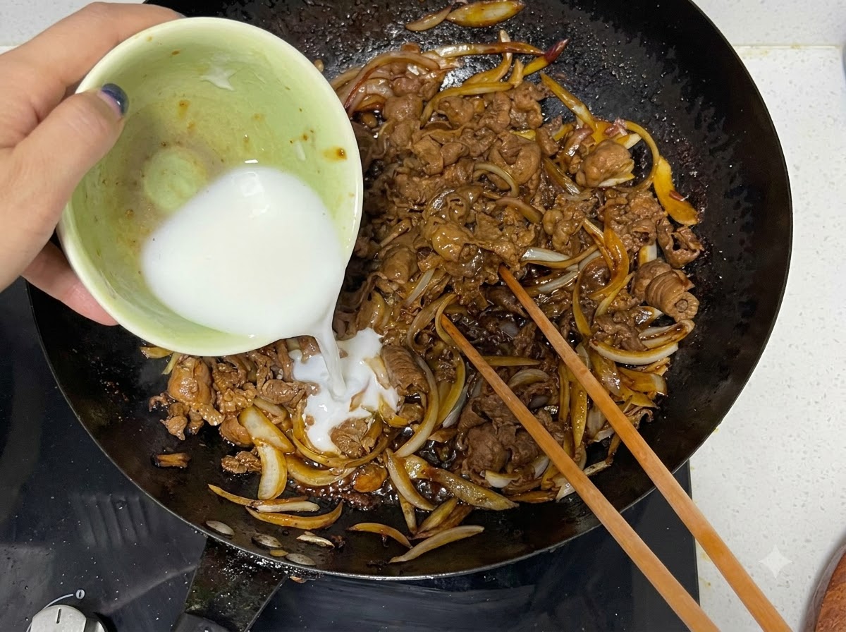 Cornstarch slurry being poured from a bowl into a wok with cooked beef and onions, alongside wooden chopsticks.