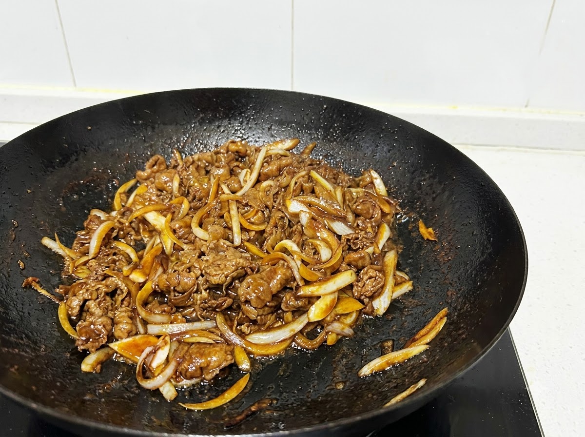 A close-up shot of beef slices and onion strips being stir-fried in a black wok with cooking utensils.