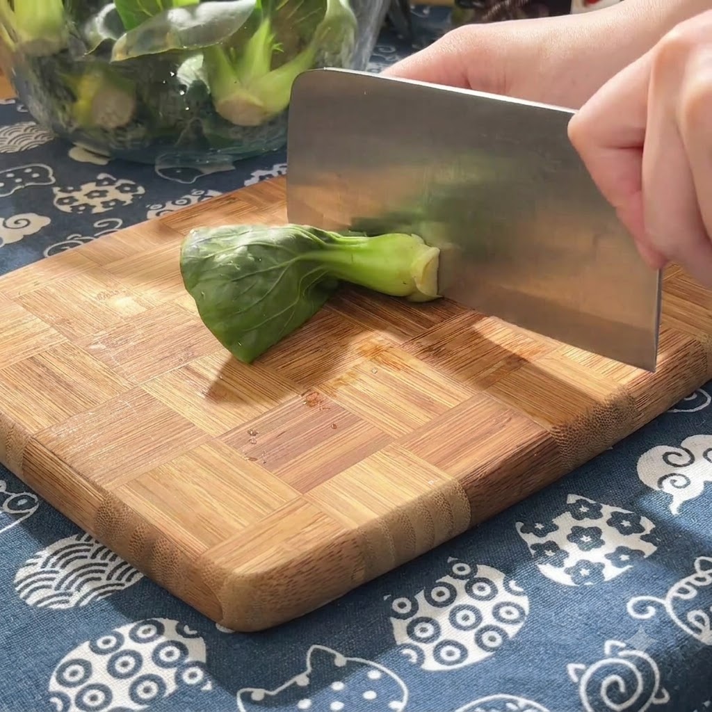 Green baby greens being sliced into small sections on a wooden chopping block using a heavy cleaver.