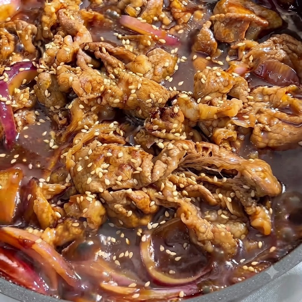 Close-up view of cooked beef and red onions simmering in a dark savory sauce, covered with white sesame seeds.
