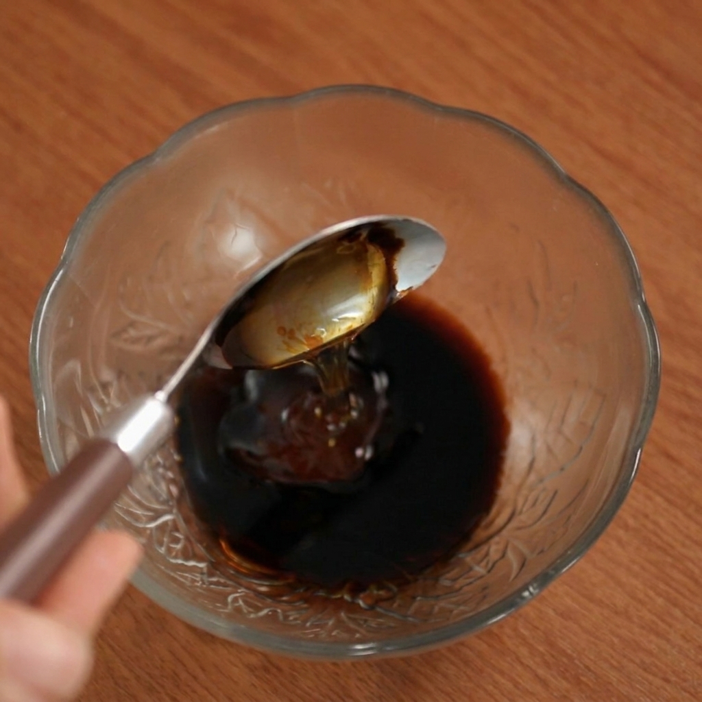 Honey being poured from a spoon into the dark sauce blend in a glass bowl.