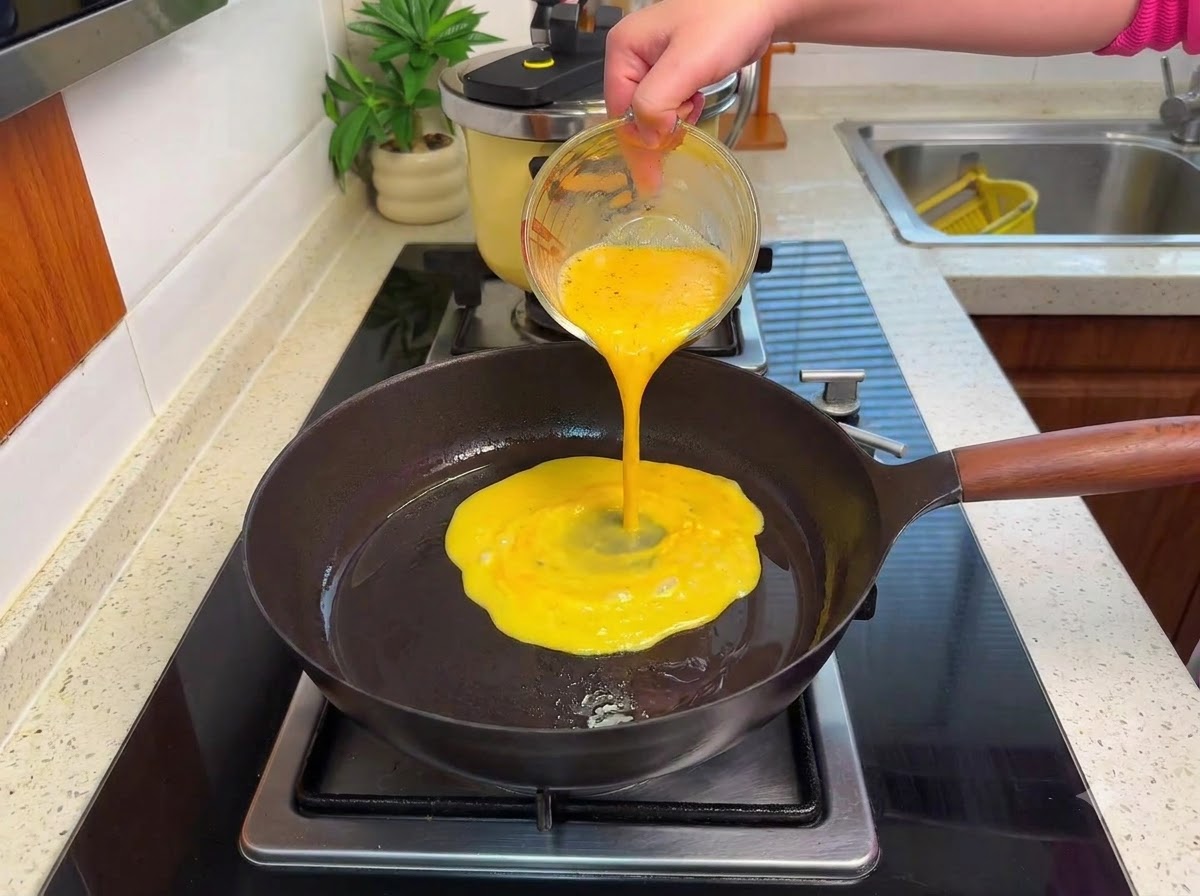Whisked eggs being poured into a hot frying pan on a stove, forming soft curds over low heat.
