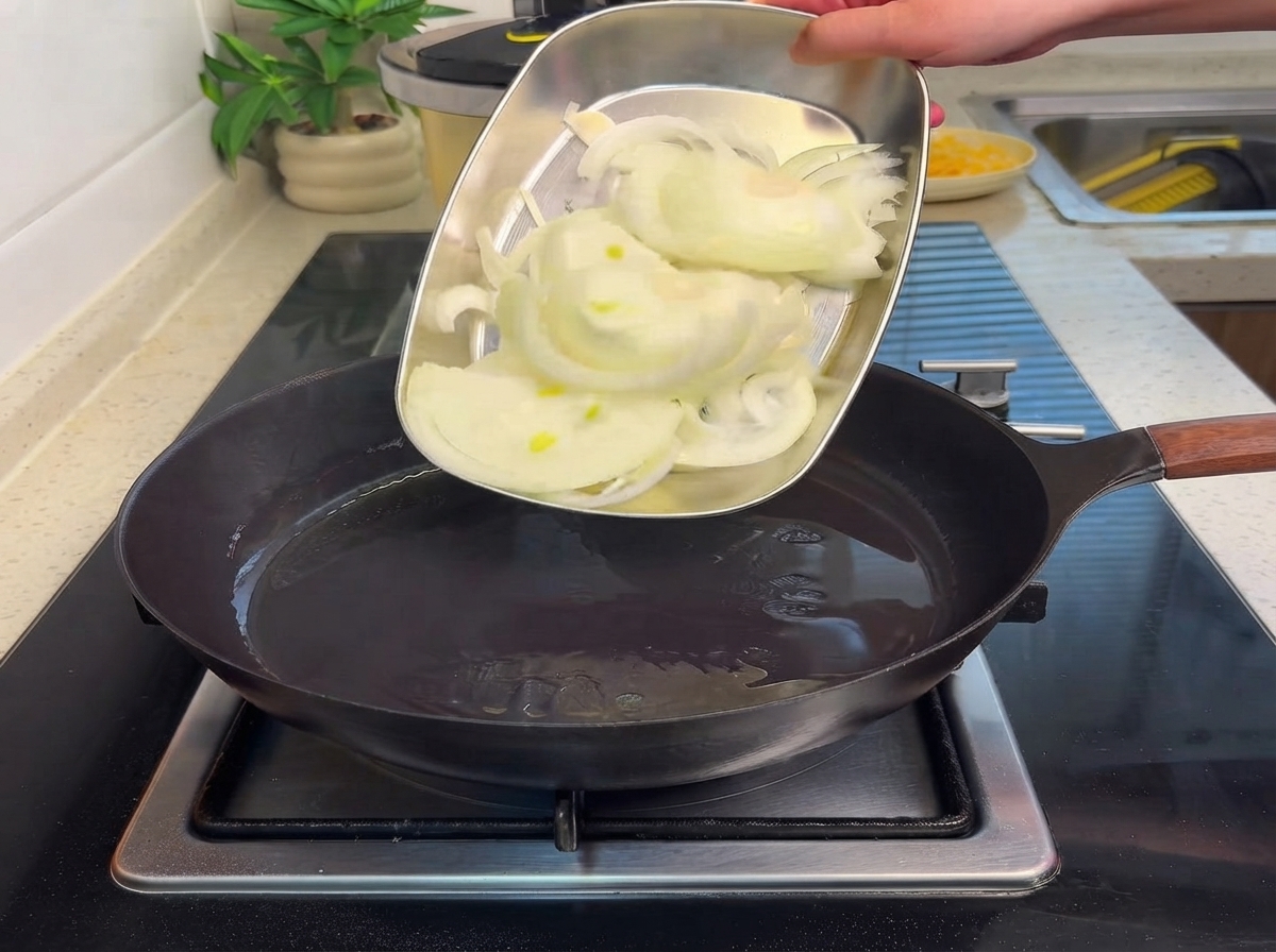 Sliced onions being slid from a metal tray into a hot frying pan with oil.