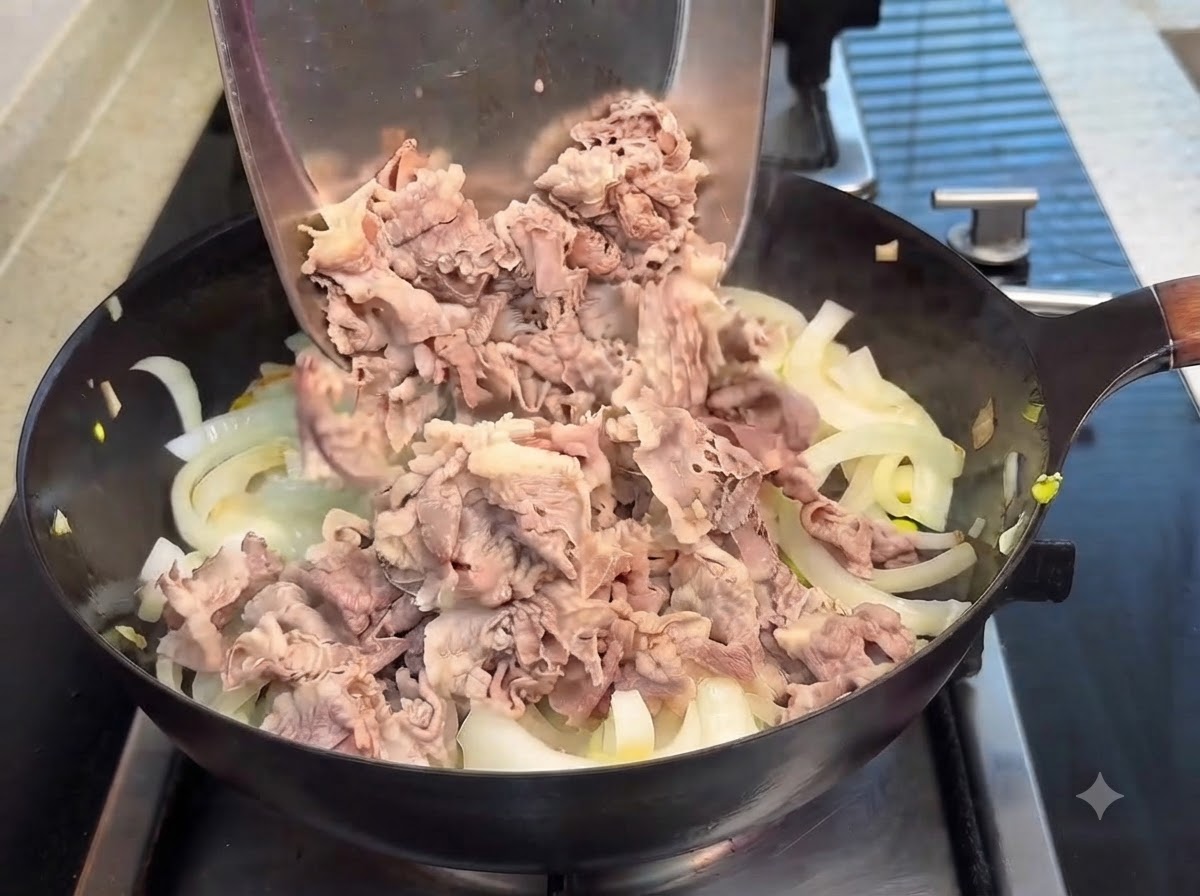 Blanched beef slices being poured from a metal spatula into a pan of translucent onions, with teriyaki sauce poured over them.