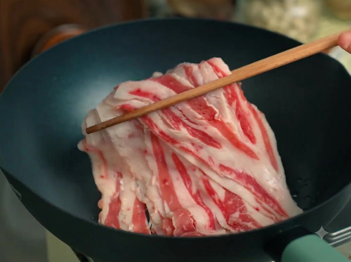 Sliced beef fat being stirred with wooden chopsticks inside a black pan.