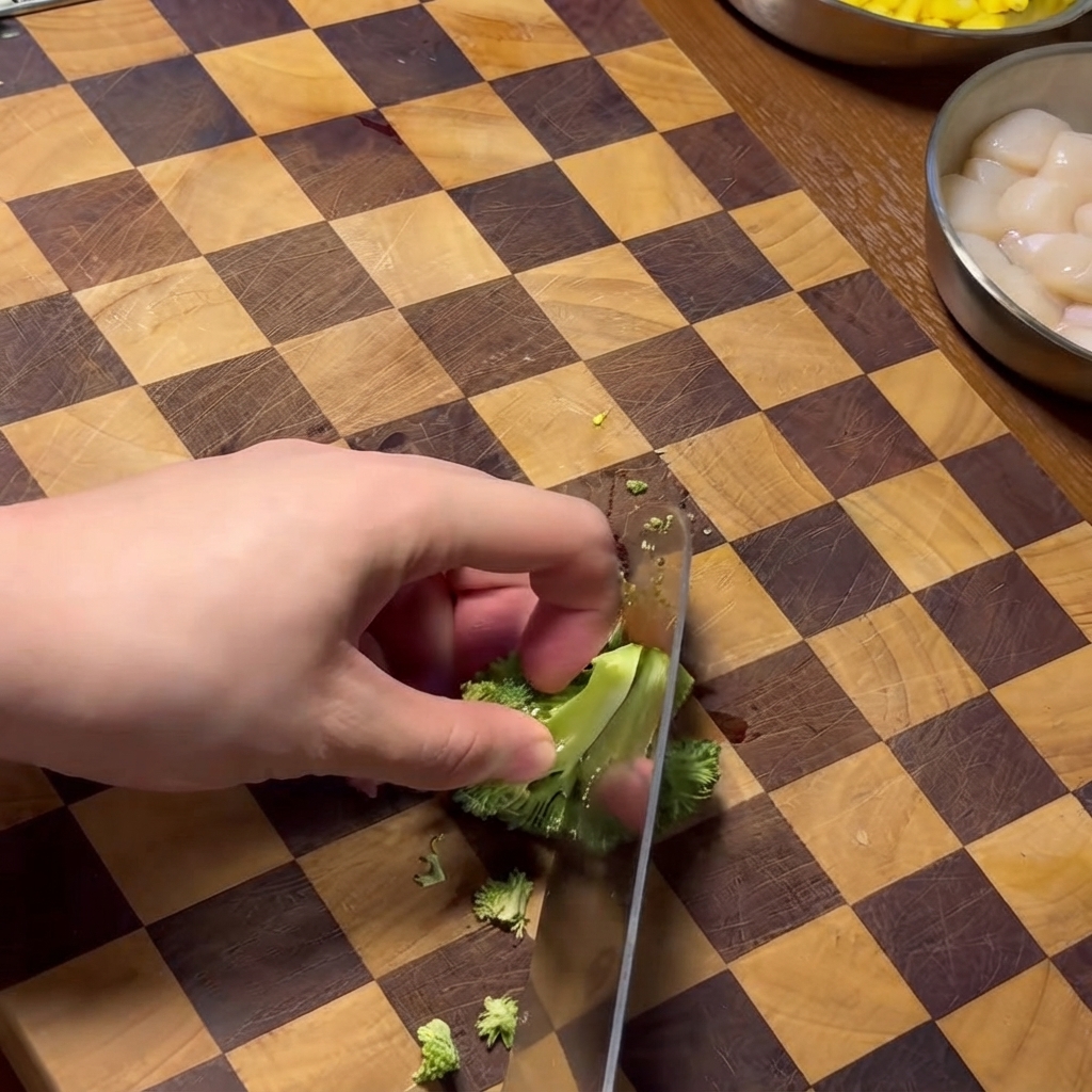 A hand using a knife to chop broccoli florets on a wooden checkered cutting board.