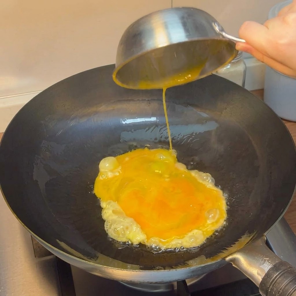 Beaten eggs being poured from a steel bowl into a heated wok.