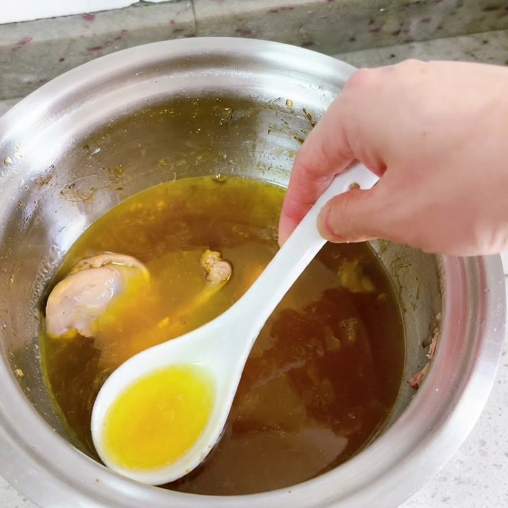 A white spoon skimming bright yellow oil from the surface of a simmering duck soup.