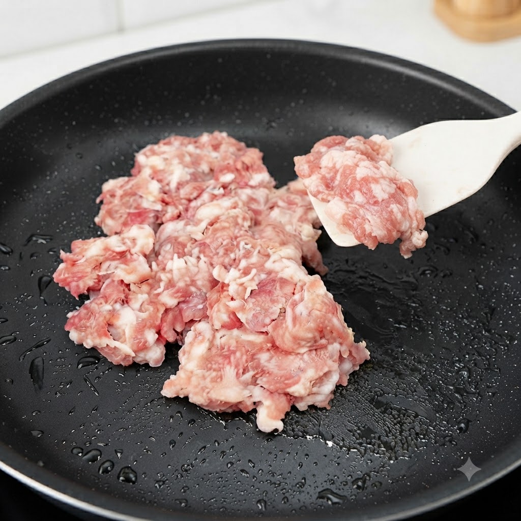 Raw minced pork being added and stir-fried in a black skillet with a silicone spatula.