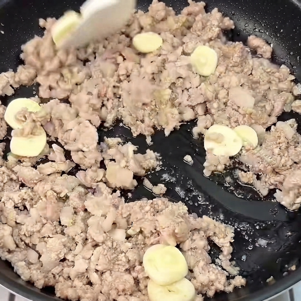 Sliced ginger and garlic being mixed and stir-fried with browned minced pork in a skillet.