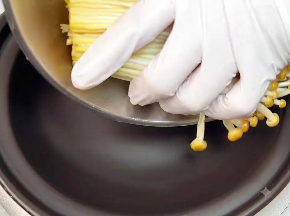 Gloved hands placing a bundle of trimmed raw enoki mushrooms into the bottom of a black clay pot.