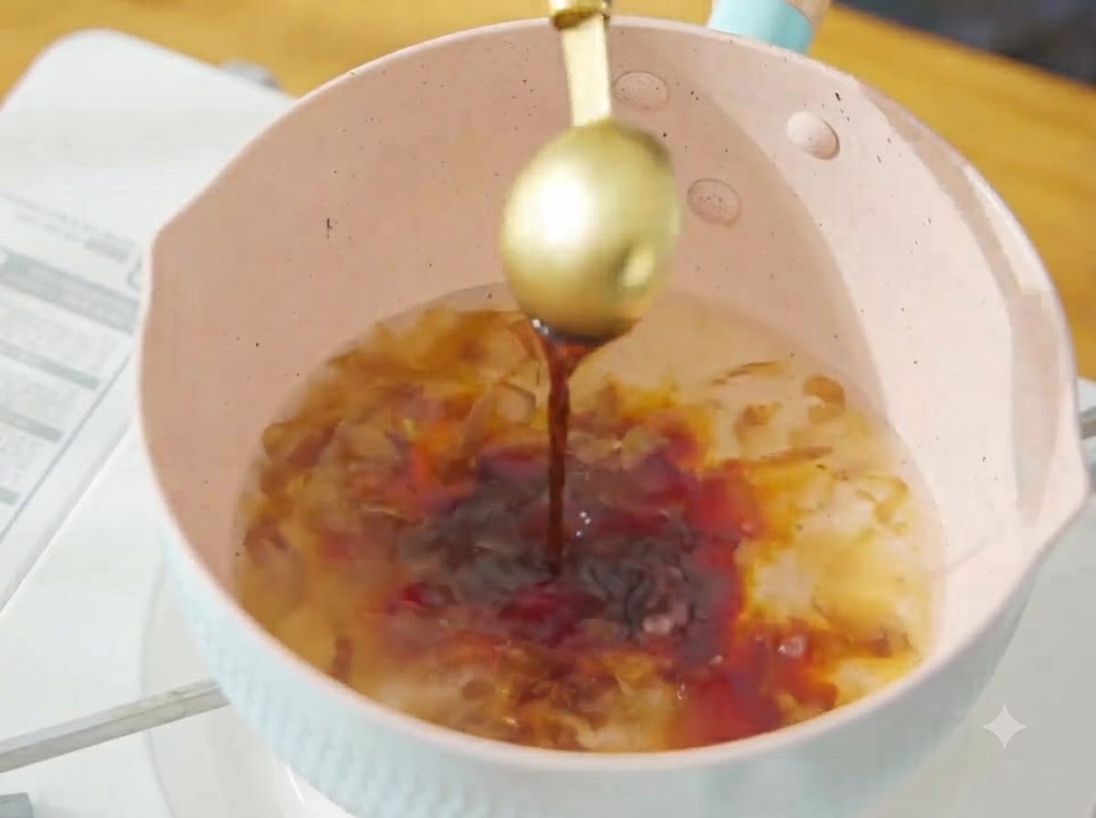 Pouring dark soy sauce from a golden spoon into the simmering bonito broth in the pink pot.