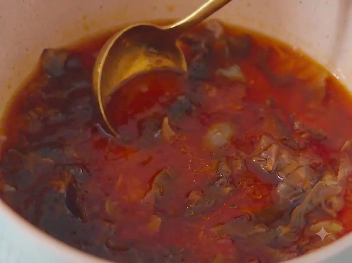 A close-up shot of a golden metal spoon stirring a reddish-brown sauce mixture with bonito flakes inside a light-colored pot.