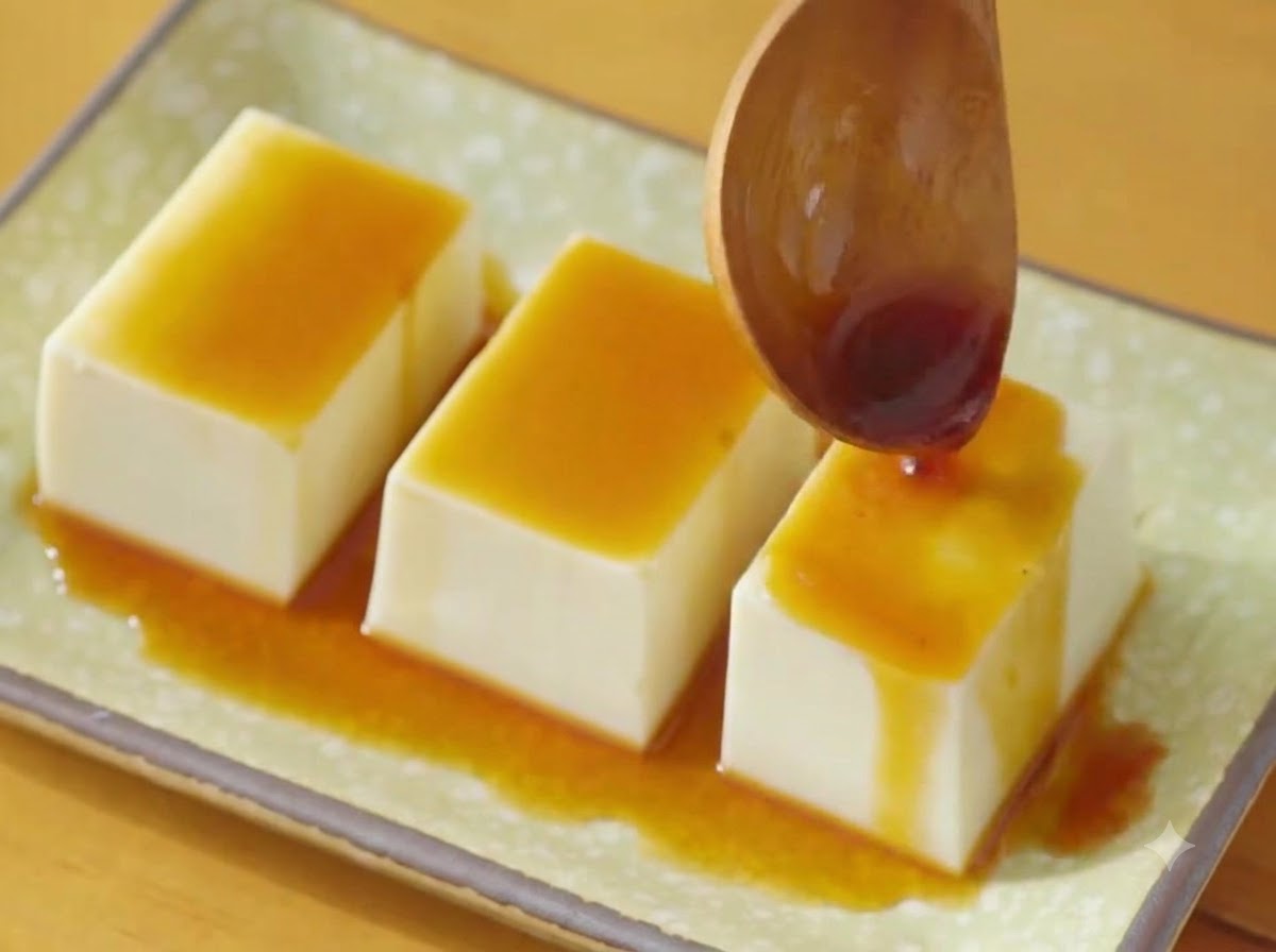 A wooden spoon pouring dark soy sauce over three rows of silken tofu on a green plate.