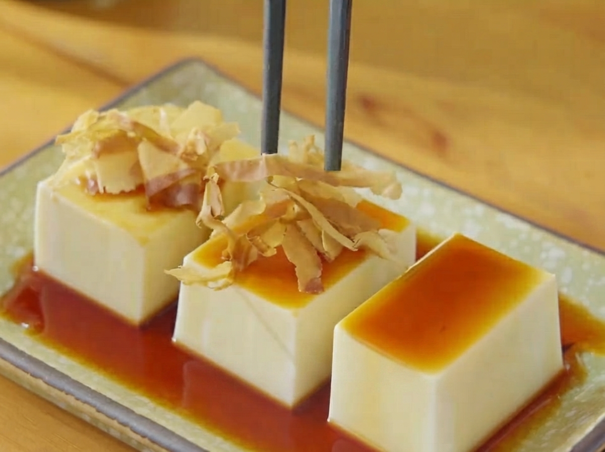 Chopsticks placing dry bonito flakes and seaweed shreds onto the soy-drizzled tofu.