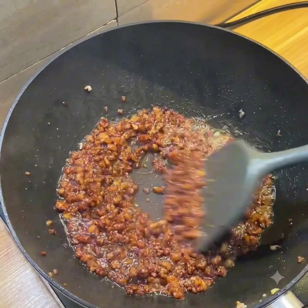 A dark wok showing rich brown minced meat sauce being stirred with a grey spatula.