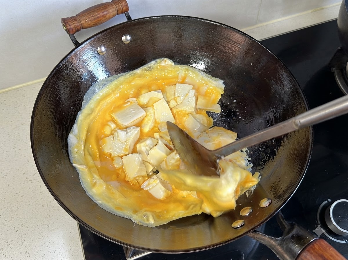 A spatula lifting and turning over the cooked layer of eggs and tofu in the wok.