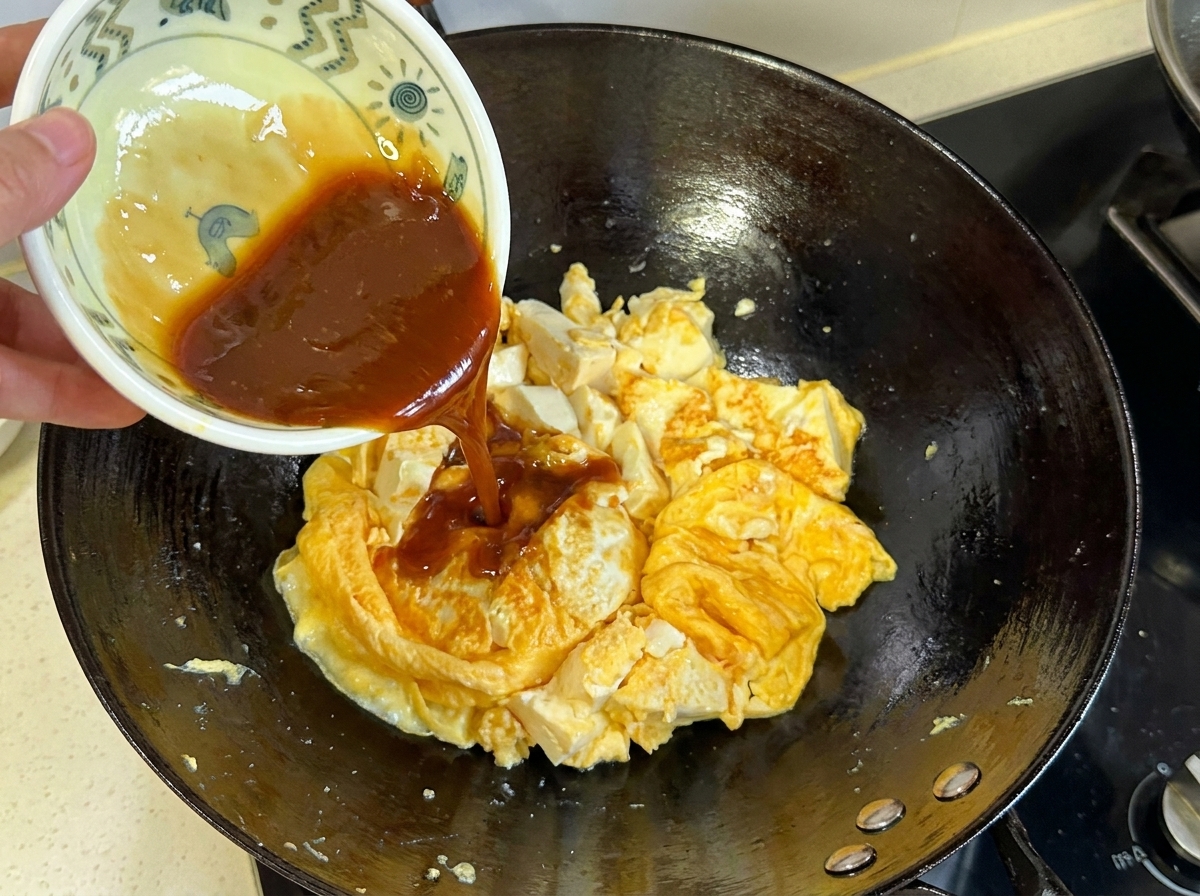Dark brown sauce being poured from a small bowl onto the cooked egg and tofu mixture in the wok.