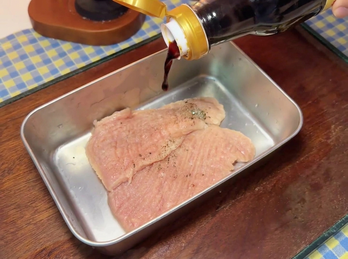 Dark soy sauce pouring from a glass bottle onto seasoned pork cutlets inside a metal tray.