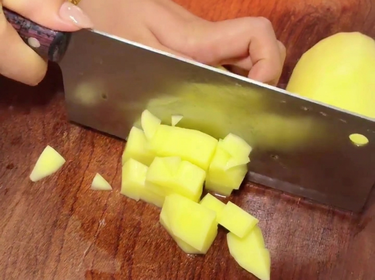A cleaver slicing through yellow potato chunks on a wooden cutting board to create small cubes.