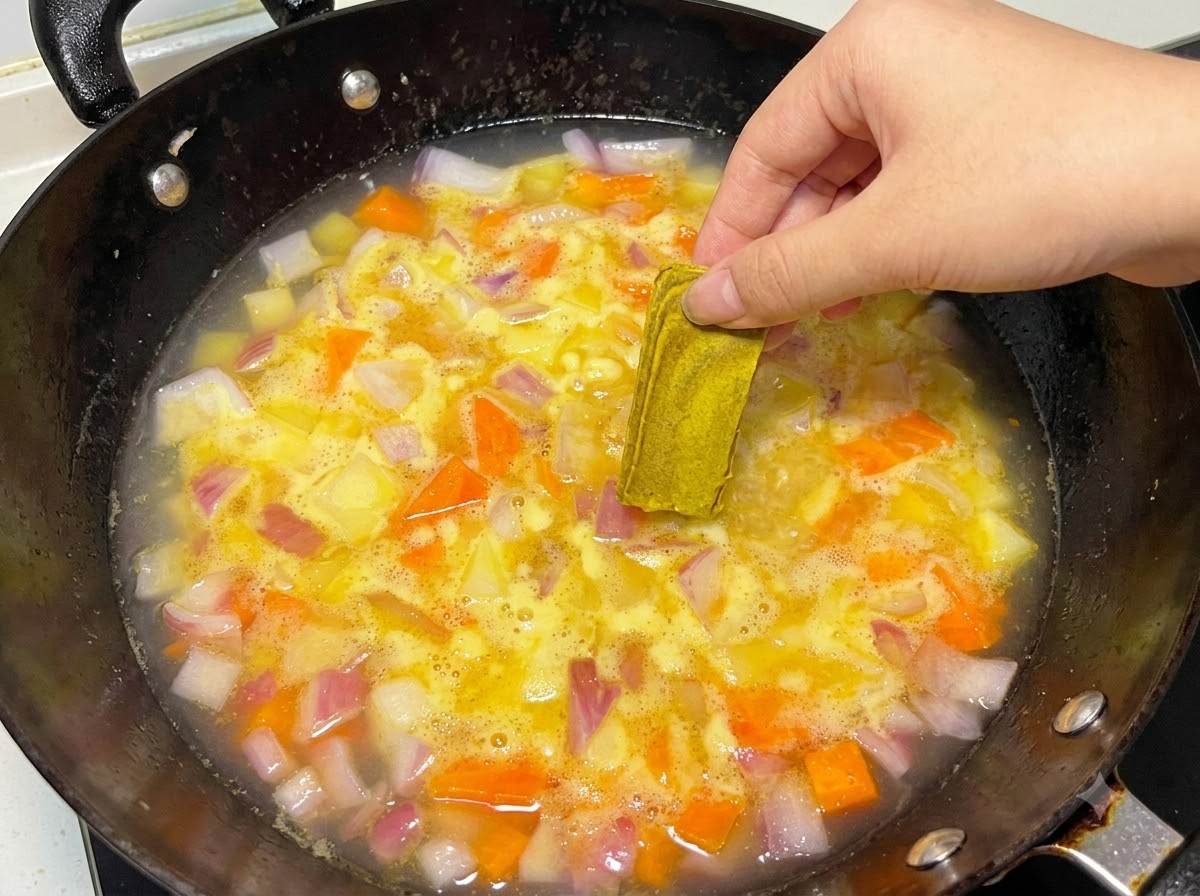 A hand placing a solid curry block into a simmering pot of water and vegetables.