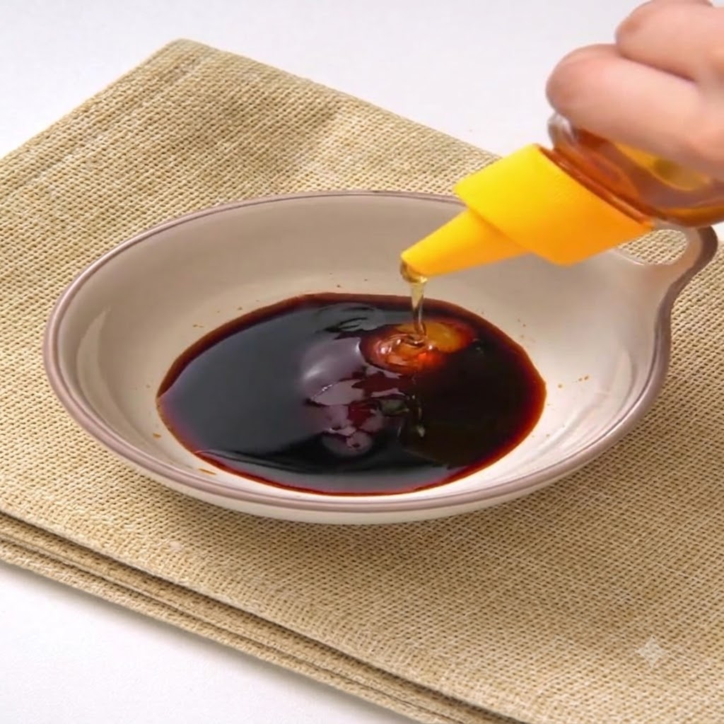 Honey being squeezed from a yellow-capped bottle into the dark sauce mixture inside a ceramic bowl.