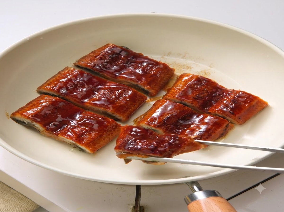 Several glazed eel slices arranged in a white pan, with metal tongs holding one piece.