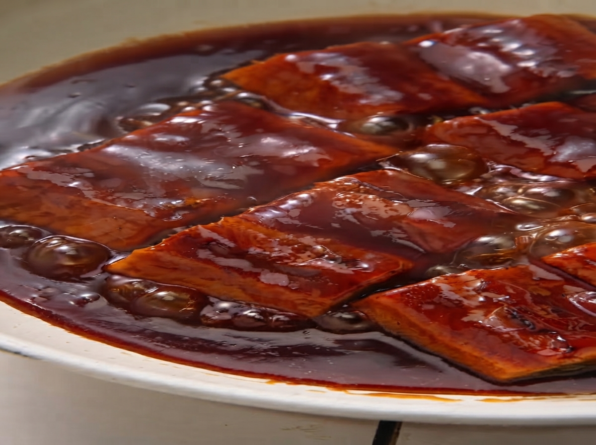 Eel pieces simmering in a bubbling, thick brown sauce inside a pan.
