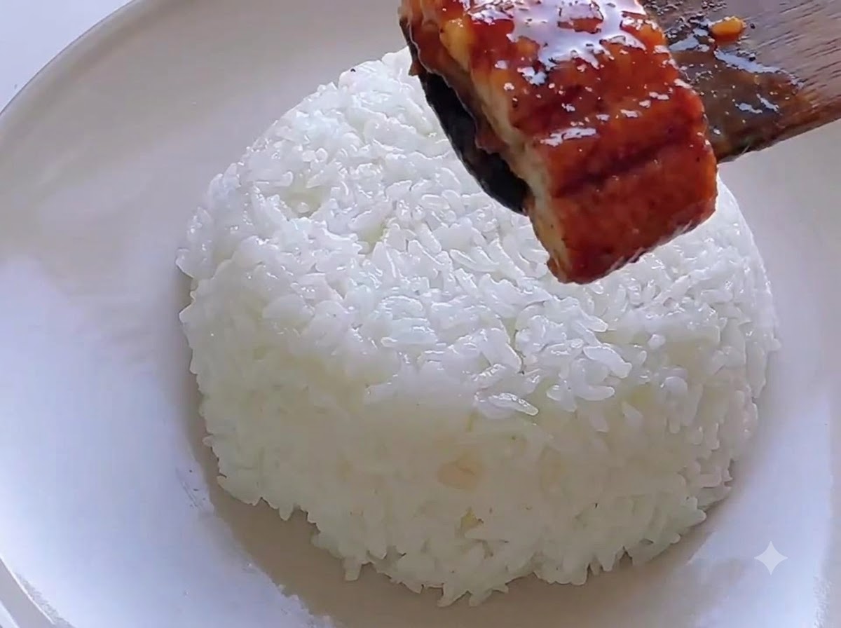 A wooden spatula placing a glazed eel piece on top of a mound of white rice.