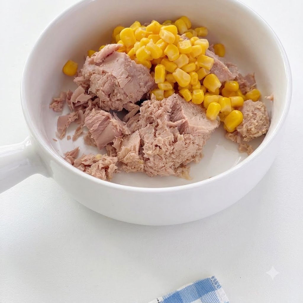 Black pepper flakes falling from a manual grinder onto a mixture of tuna and yellow corn kernels.