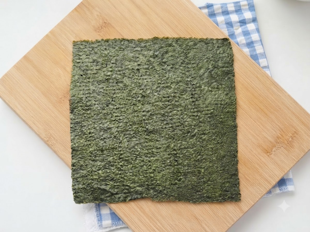 A full sheet of green sushi nori resting on a wooden cutting board with a blue checkered cloth underneath.