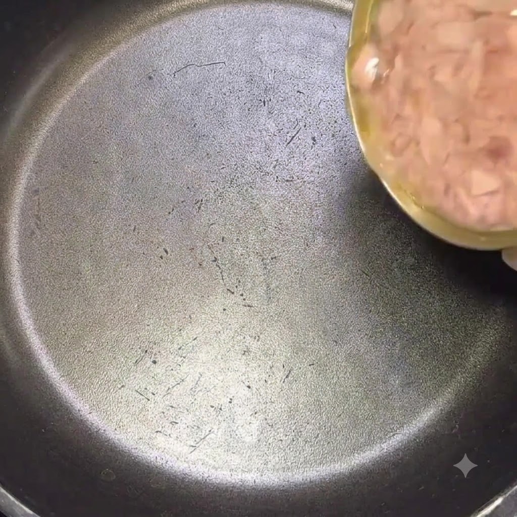 A large can of tuna being poured with its oil into a black frying pan.
