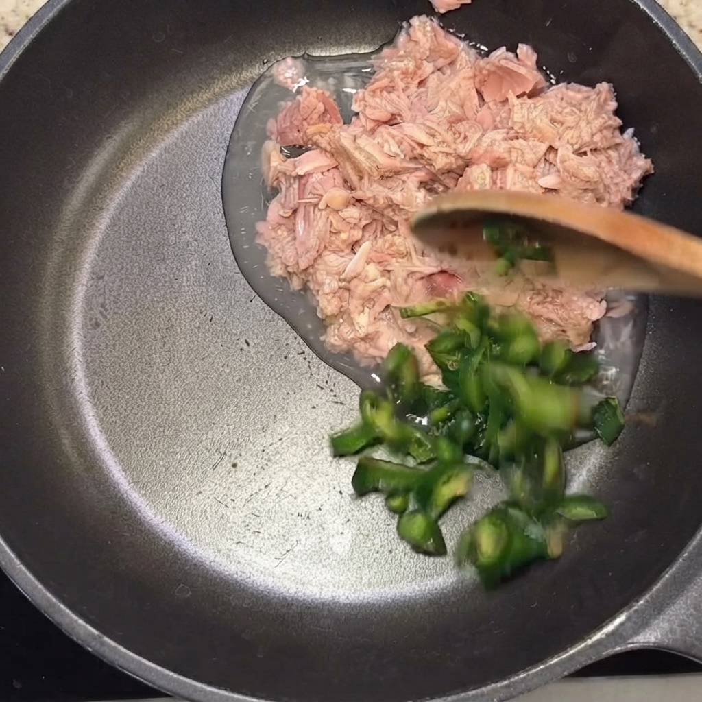 Sliced green chili peppers being added into the frying pan with the tuna using a wooden spoon.