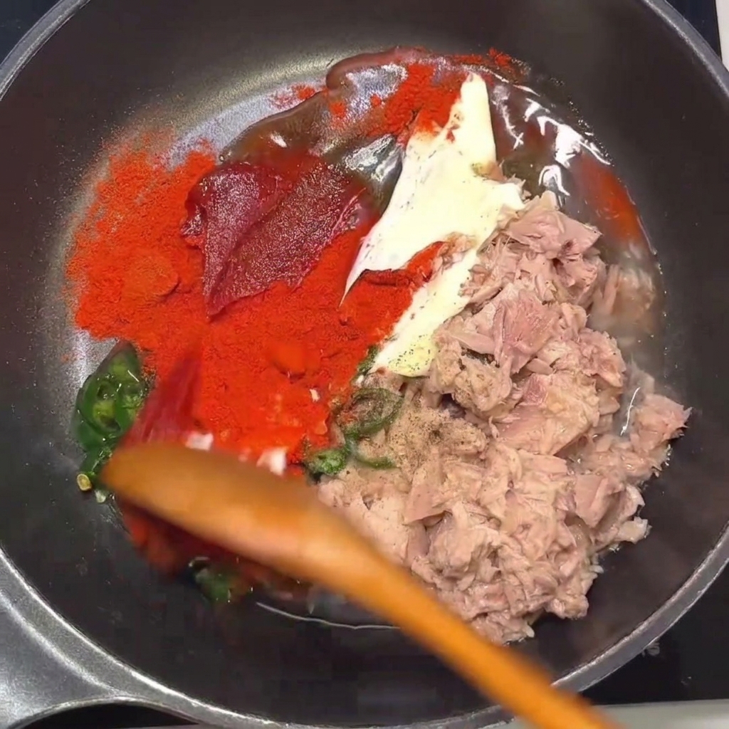A wooden spoon mixing the tuna, chili, and seasonings together in a frying pan.