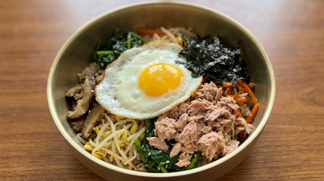 A colorful bowl of tuna bibimbap featuring seasoned rice, fresh vegetables, mushrooms, seaweed flakes, canned tuna, and a sunny-side-up egg.