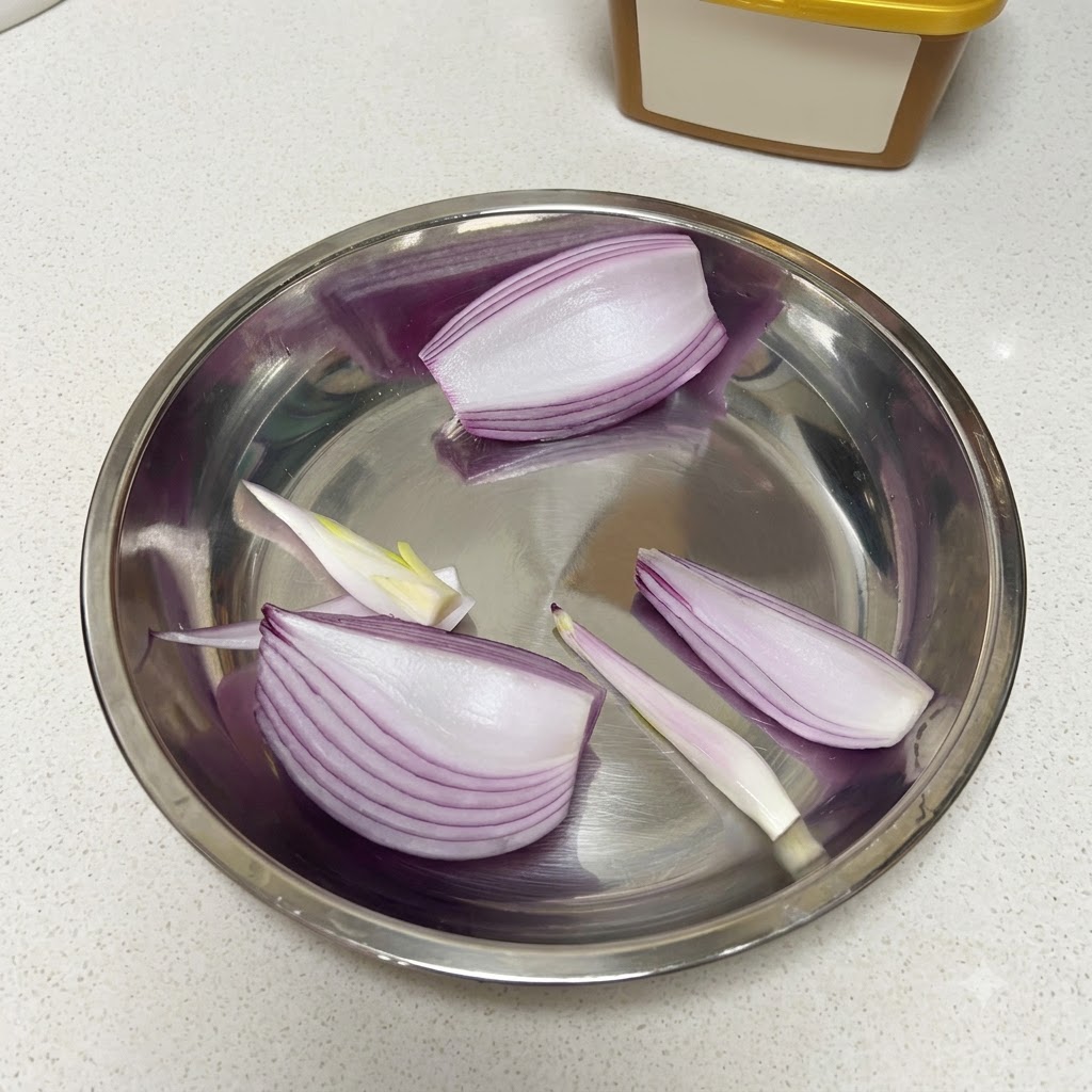Cut red onion, half an apple, and peeled raw garlic cloves arranged inside a metal mixing bowl.