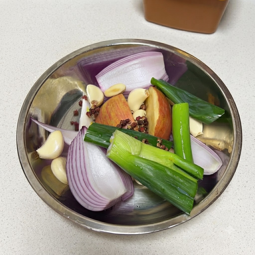 Fresh green onions being cut into a metal bowl filled with red onion, apple, garlic cloves, peppercorns, and a bay leaf.