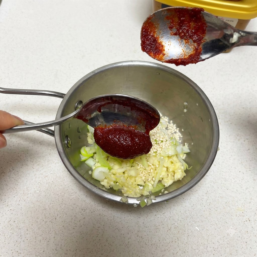 A spoonful of Korean chili paste held over a pan with garlic and sesame seeds.