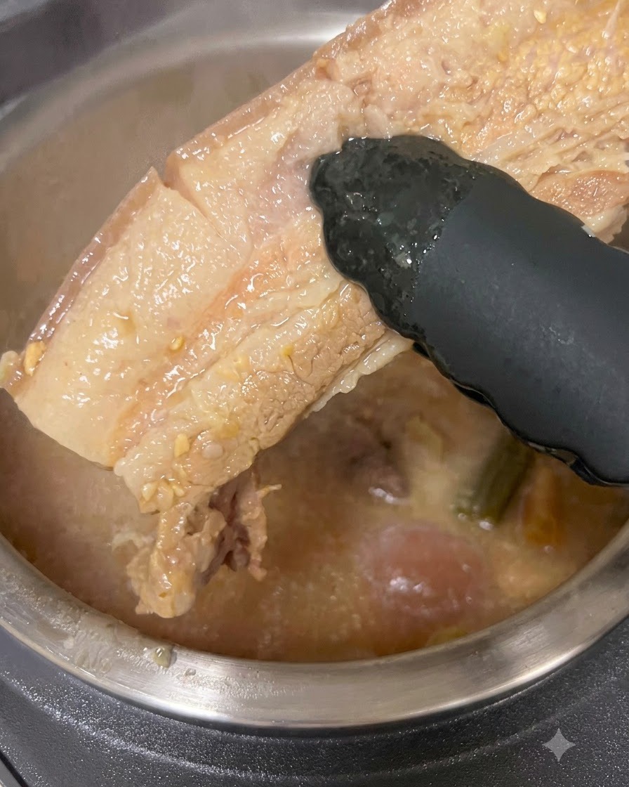 A thick piece of cooked pork belly being lifted out of a pot of broth with black tongs.