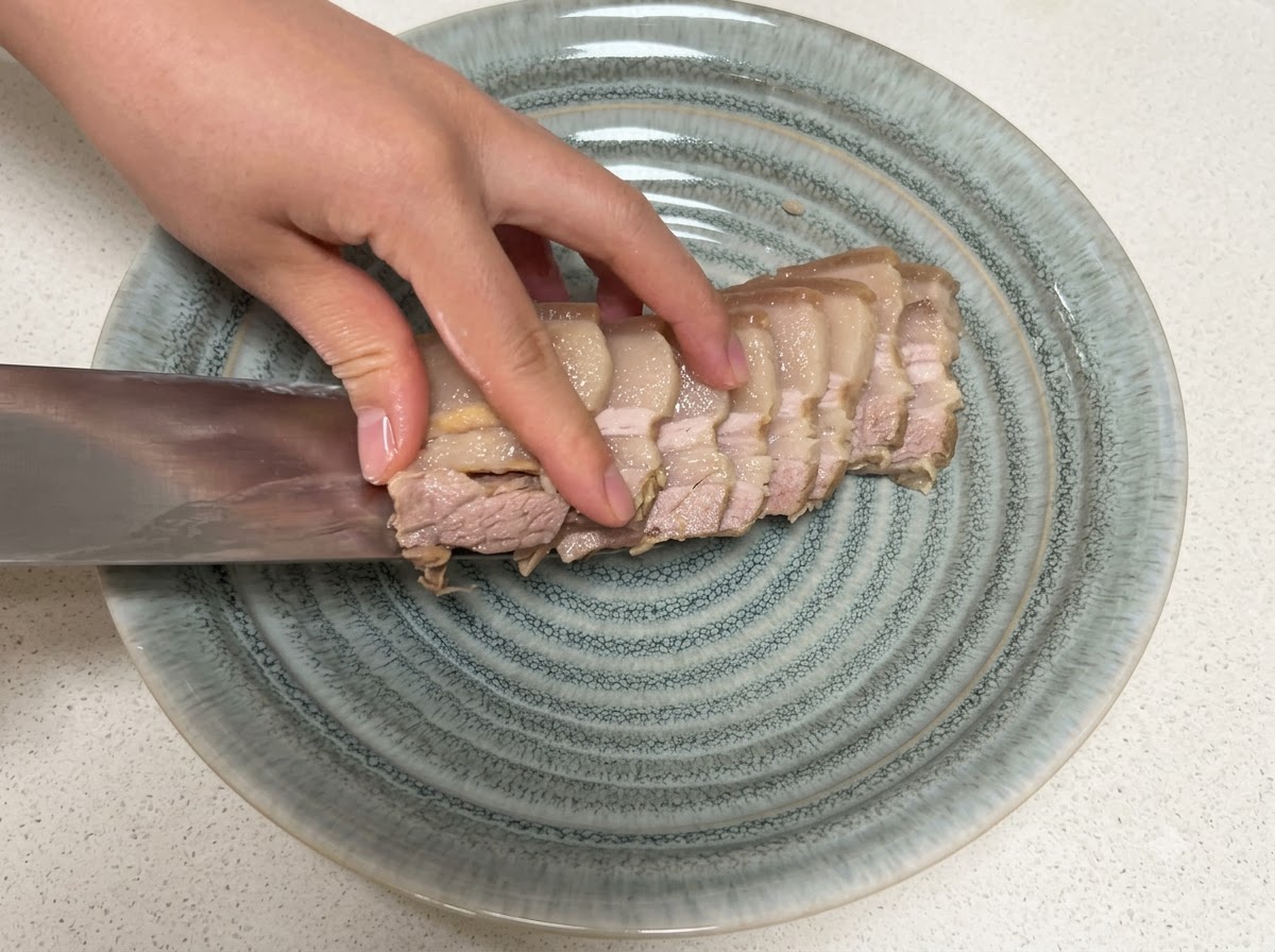 Slices of cooked pork belly being transferred onto a textured grey plate with a knife.