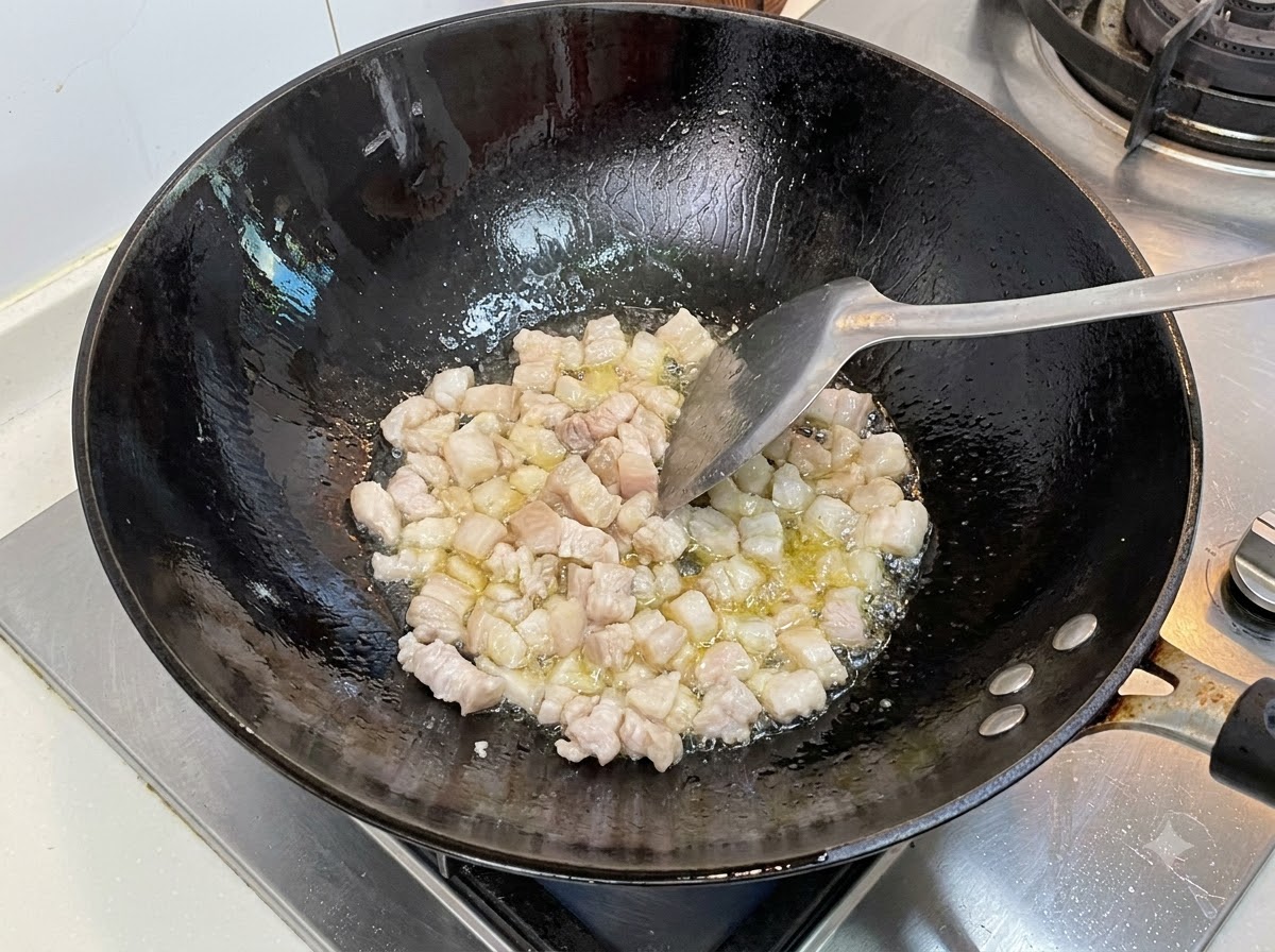 Fatty pork cubes frying in a hot black wok with cooking oil being stirred with a spatula.