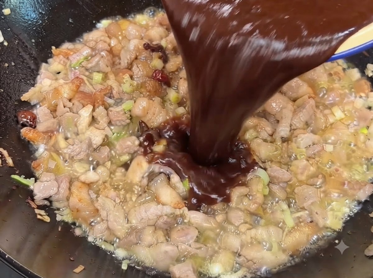 Dark soybean and sweet bean paste sauce mixture being poured into the wok over the cooked pork.