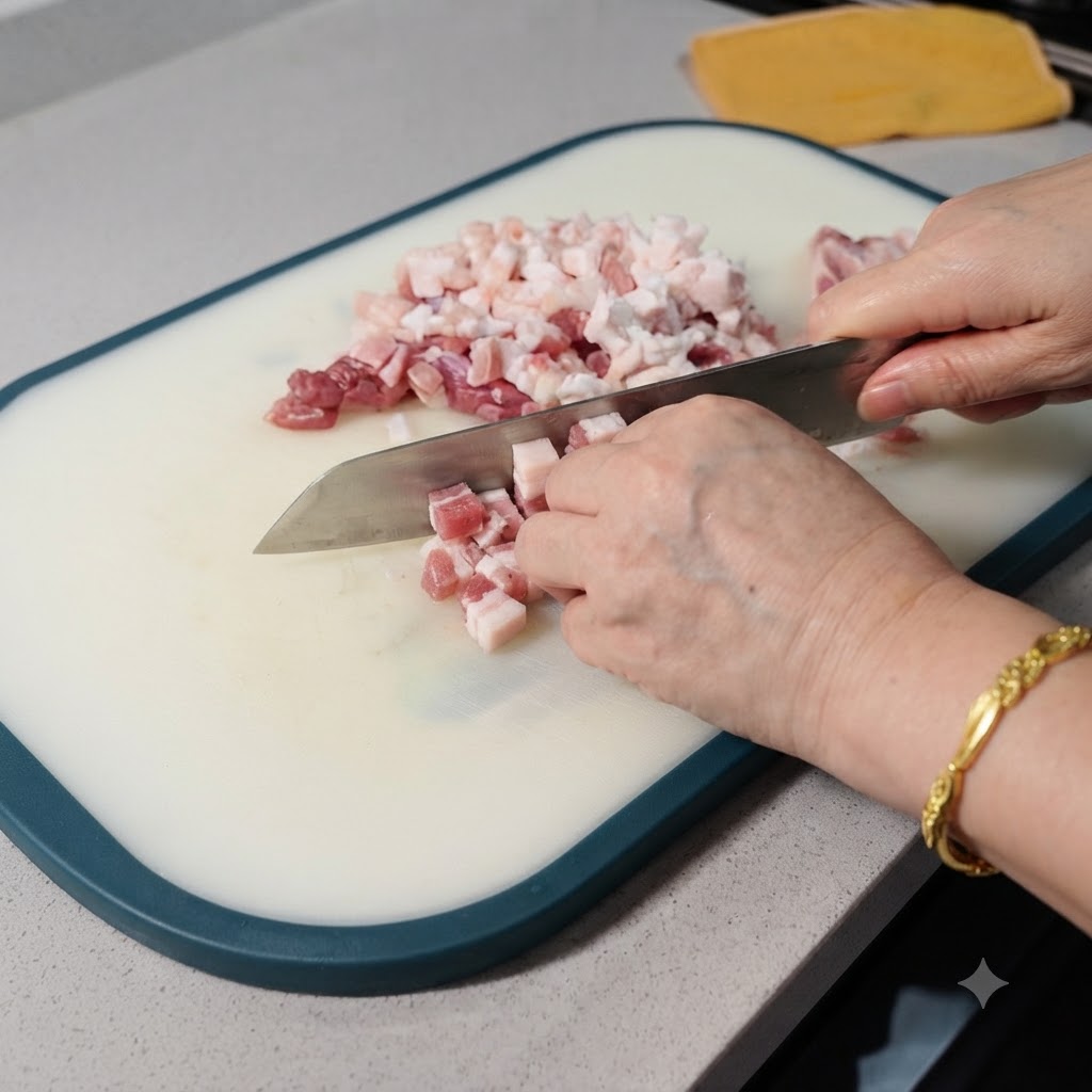 A close-up view of raw pork belly being diced into small cubes with a cleaver on a white cutting board.