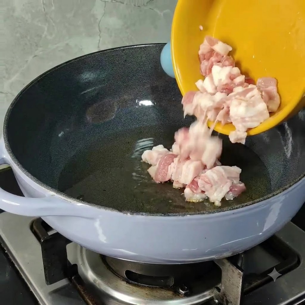 Diced raw pork belly pieces sliding from a yellow bowl into a heated pan with cooking oil.