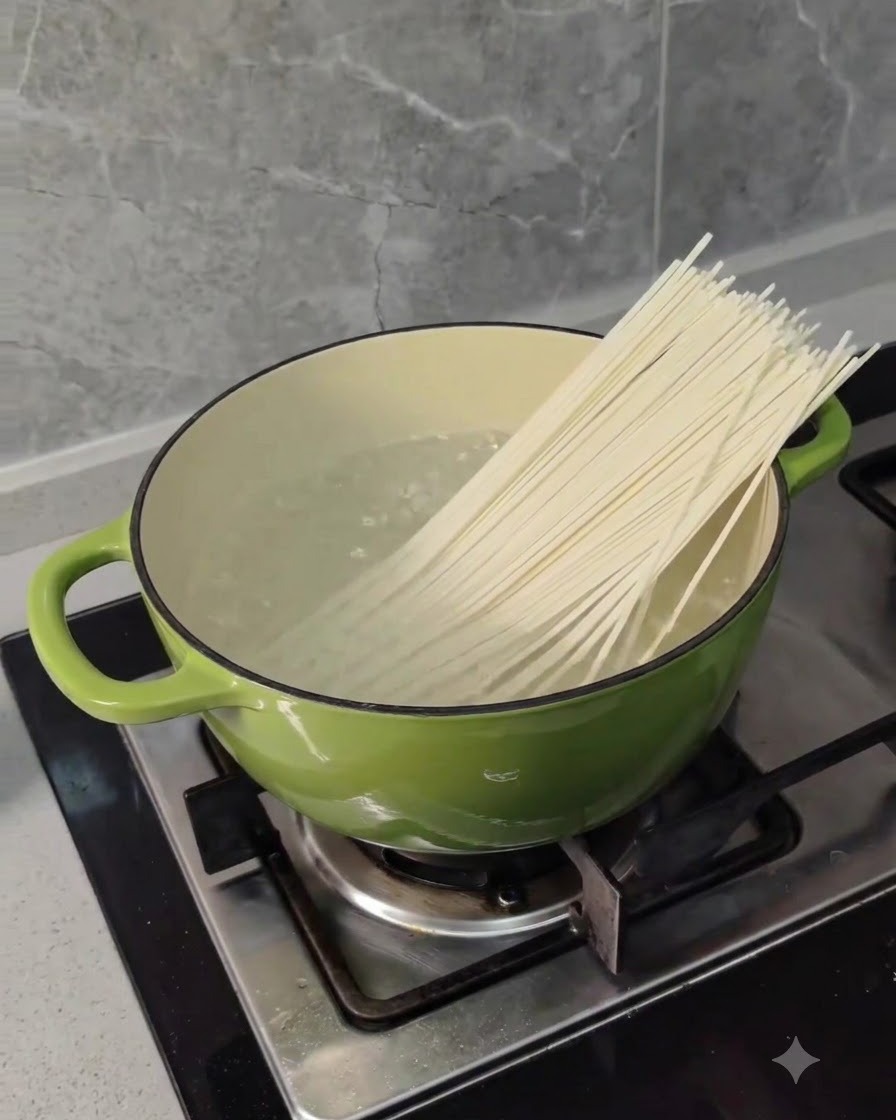 Dry long wheat noodles being placed into a green pot of boiling water on a stove.