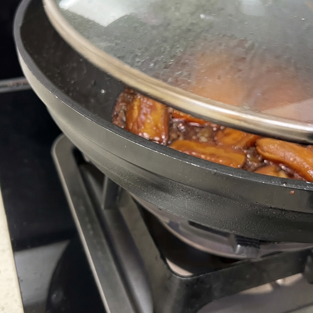 Close-up of a wok with the lid partially lifted, showing pork ribs simmering in a glossy brown reduced sauce.