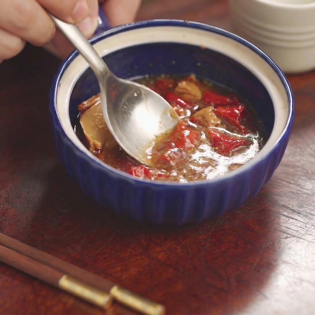 A spoon mashing and mixing red bean curd and seasonings inside a small blue ceramic bowl.