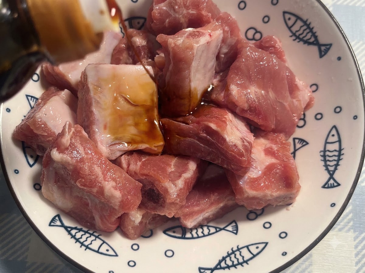 Pork ribs in a white patterned bowl being seasoned with dark soy sauce.