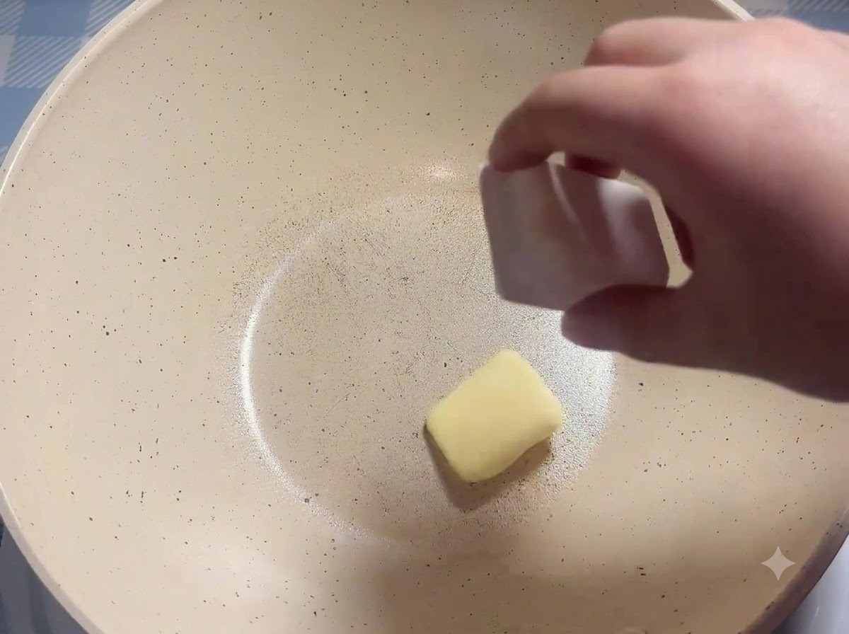A block of butter being placed into a heated white frying pan.