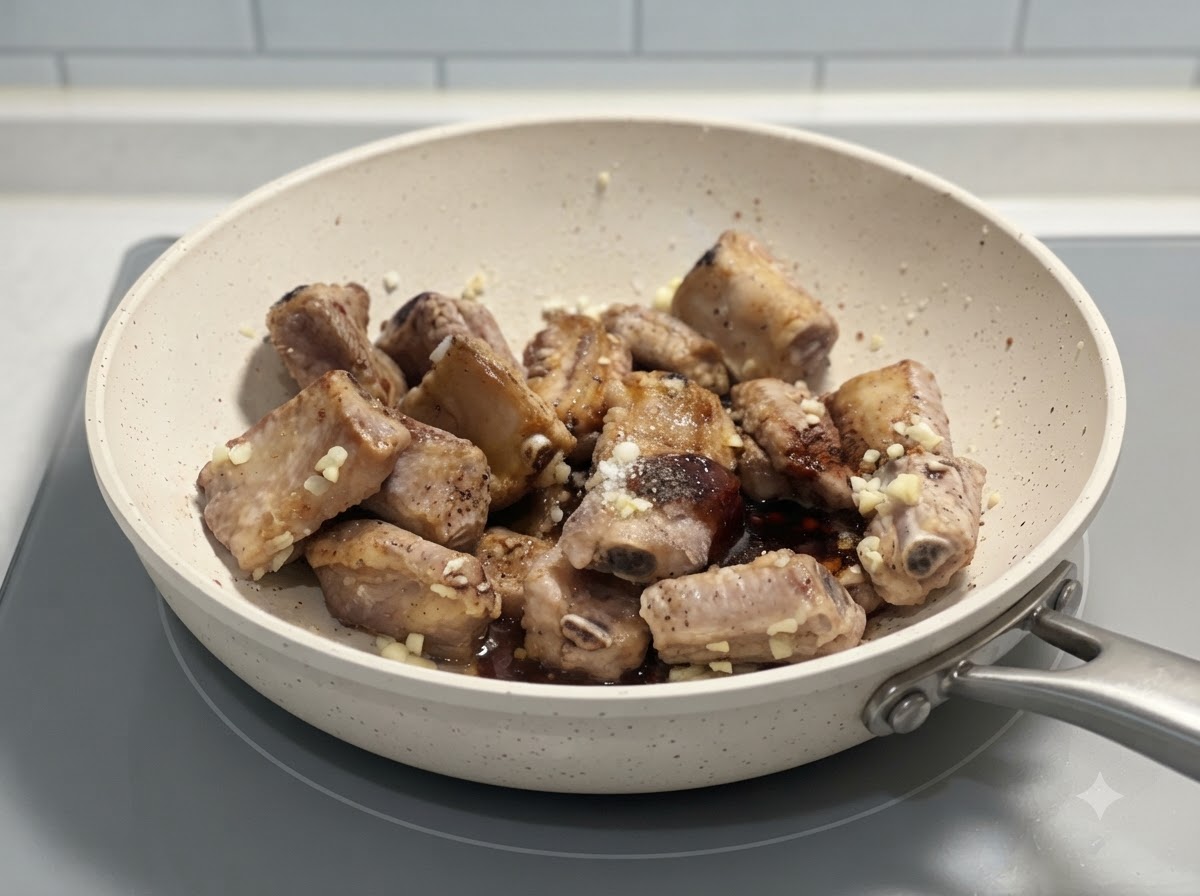 Pork ribs being tossed together with seasonings and minced garlic in a white pan on a stove.